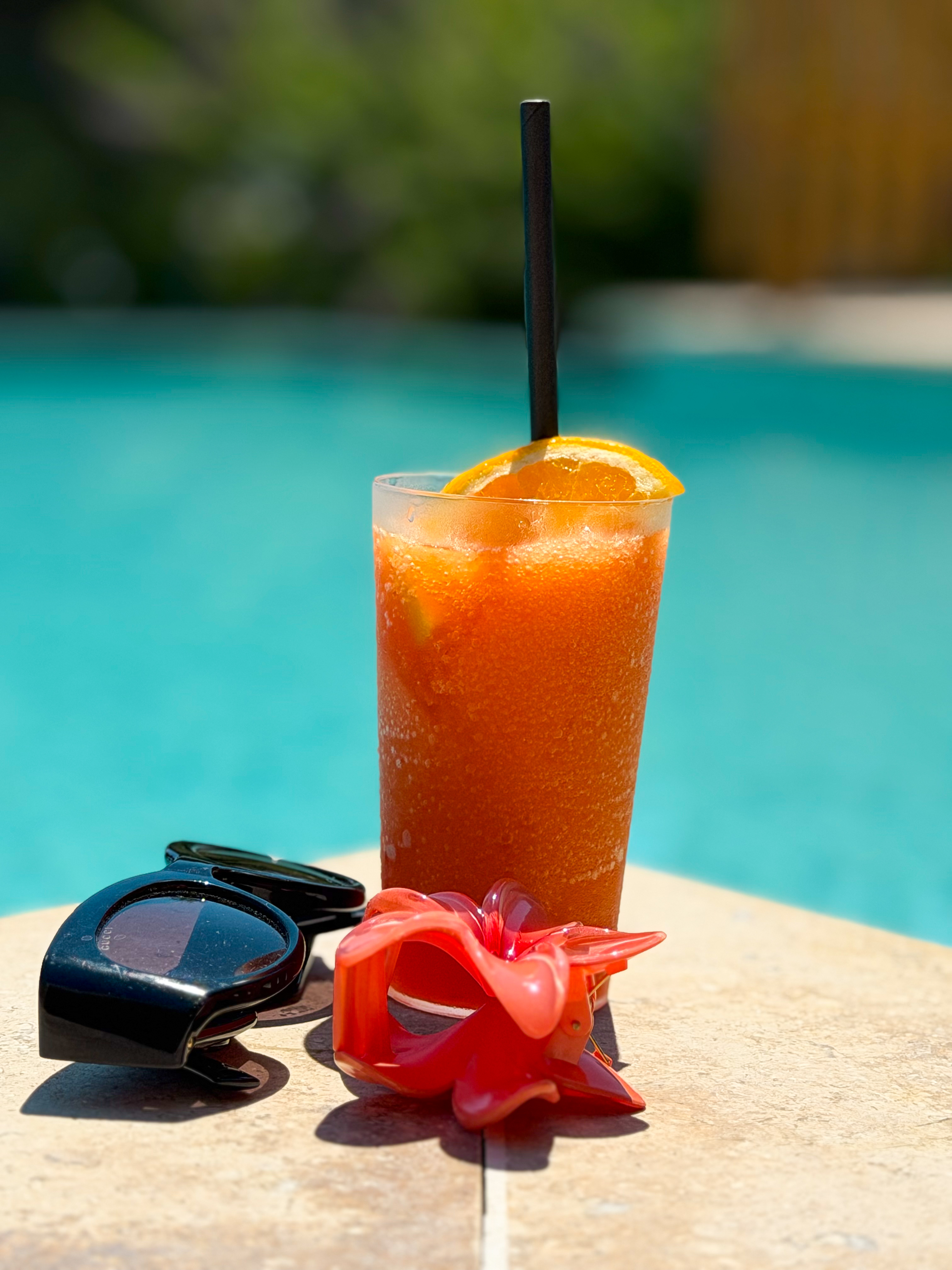 An orange frozen drink, black sunglasses and a red flower hair clip on a stone edge beside a turquoise swimming pool.