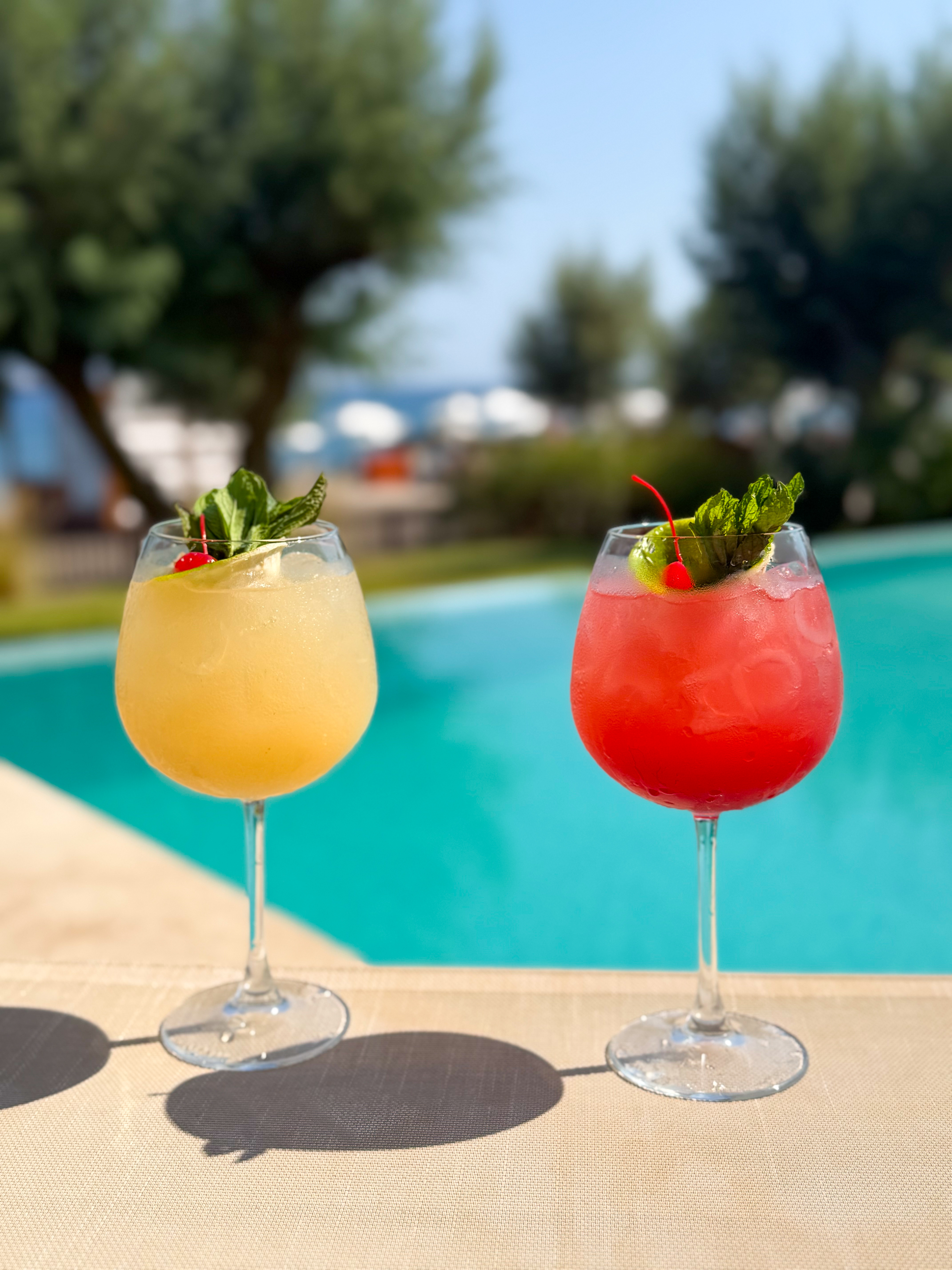 Two colourful iced cocktails with mint garnishes sitting poolside under bright afternoon sunlight.