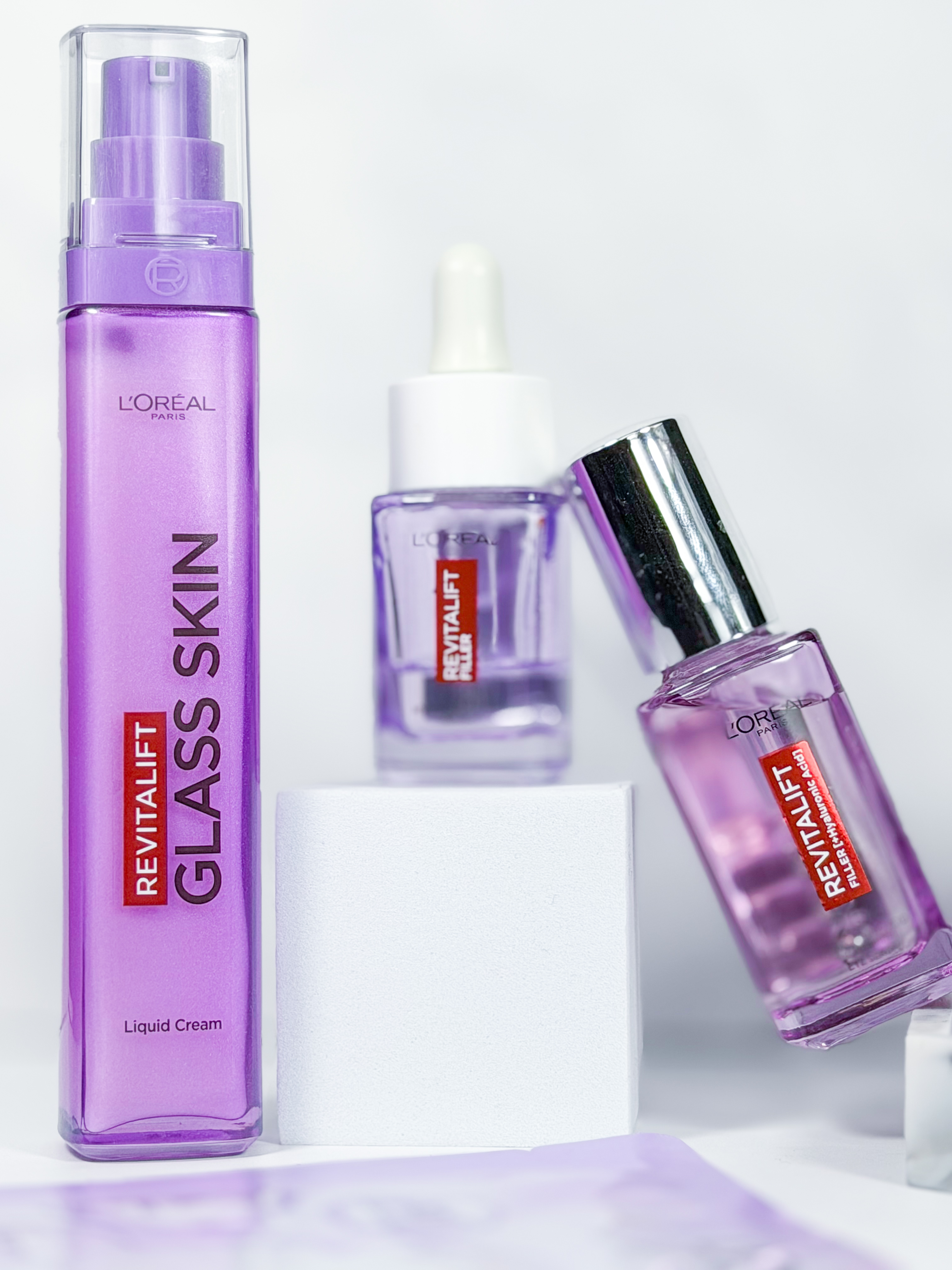 Three purple glass skincare bottles arranged on white textured blocks against a bright, minimal background.