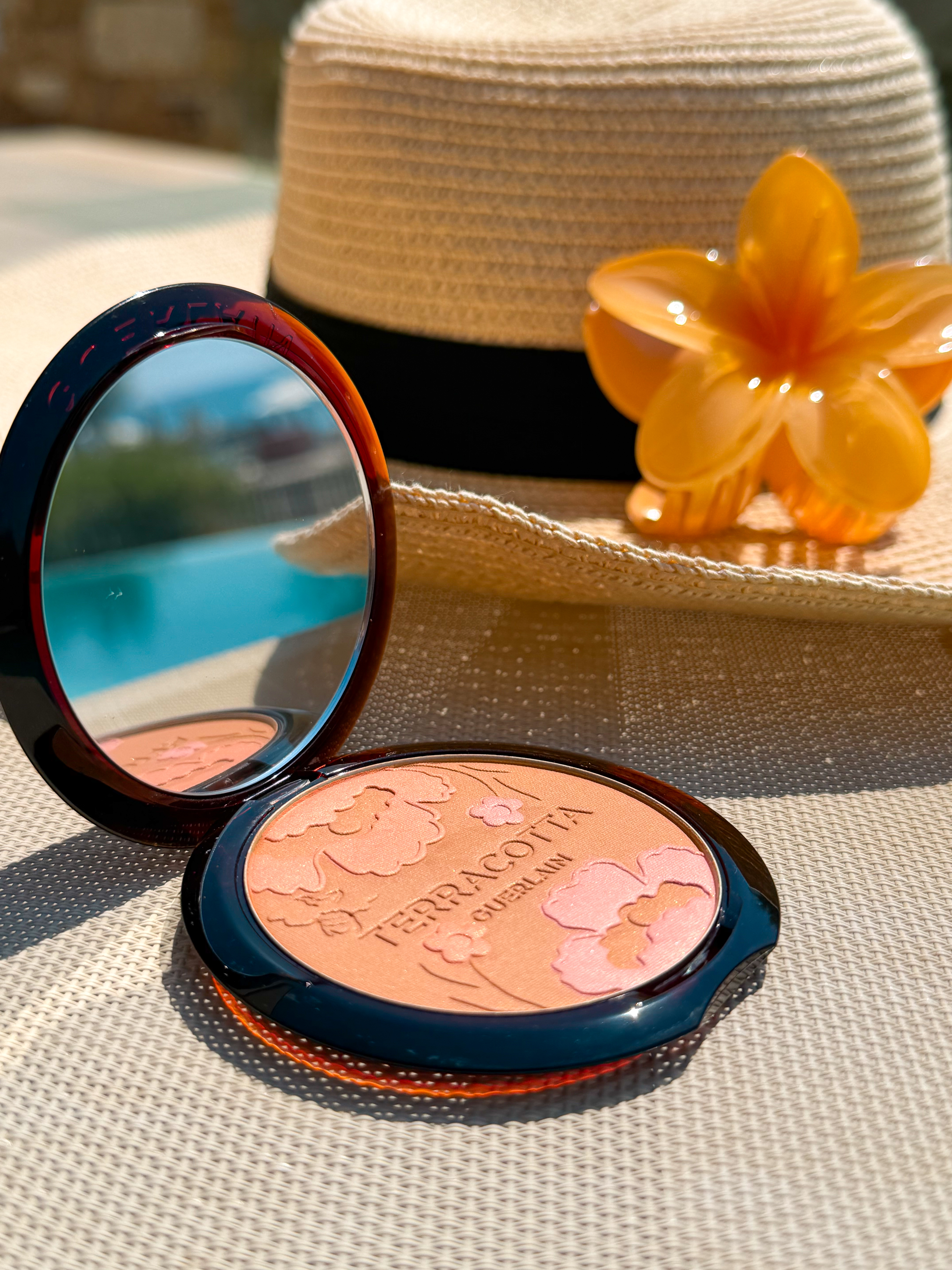 An open bronzer compact with embossed floral patterns sits on a woven surface beside a straw hat and orange flower clip.