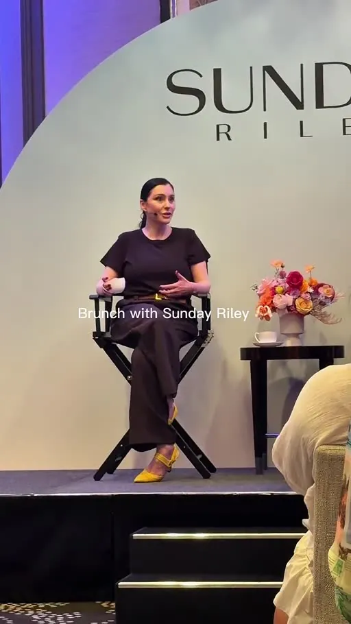 A woman in black seated on a director's chair beside colourful flowers on a stage with soft neutral tones.