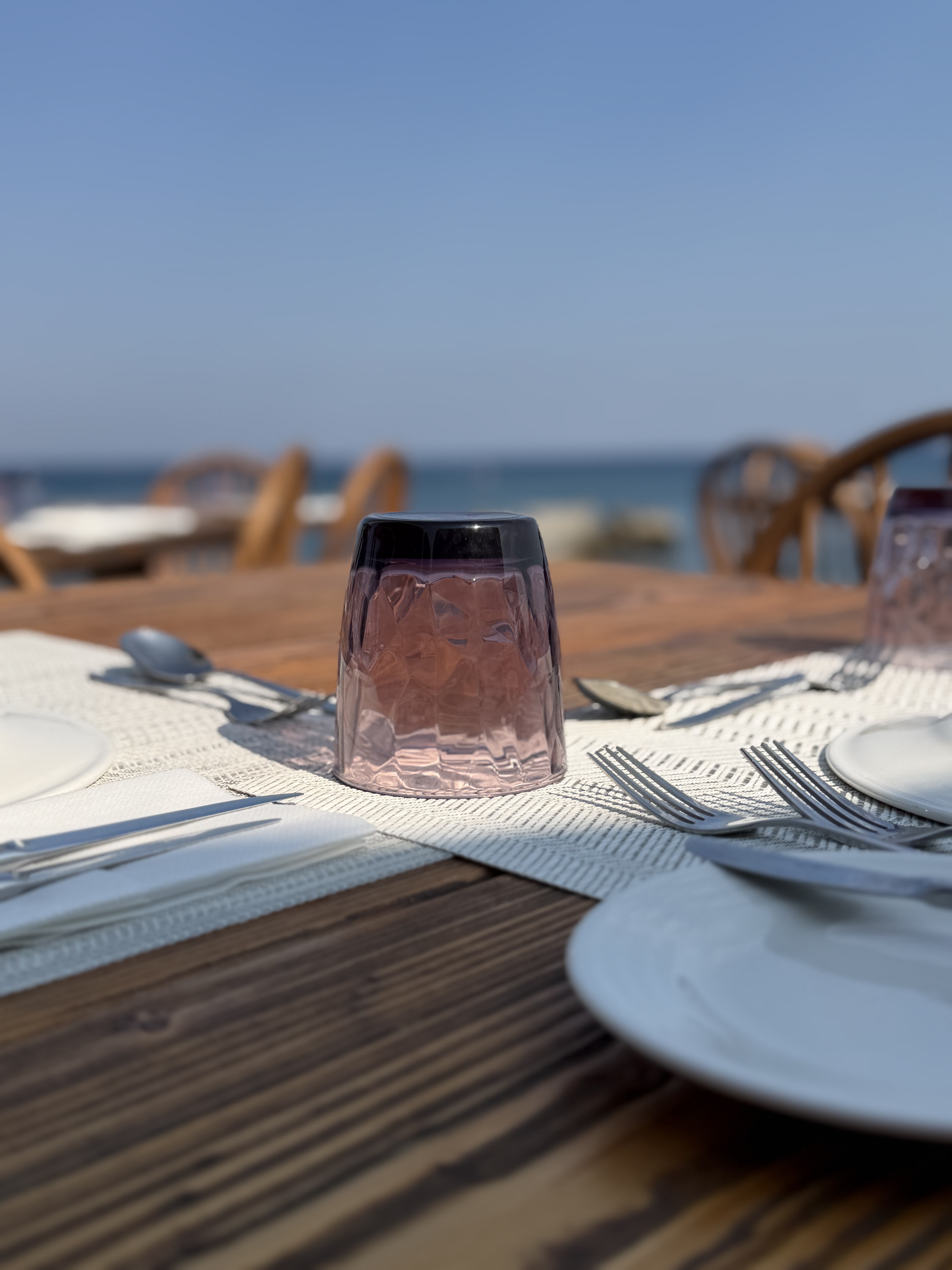Textured purple glass and silver cutlery on a wooden table overlooking the blue sea in bright summer light.