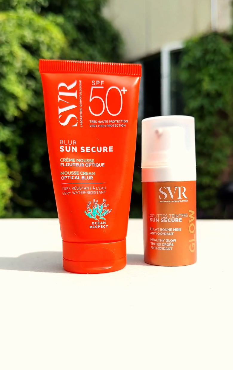 Two bright orange skincare tubes standing on a white ledge in natural light with a blurred green garden background.