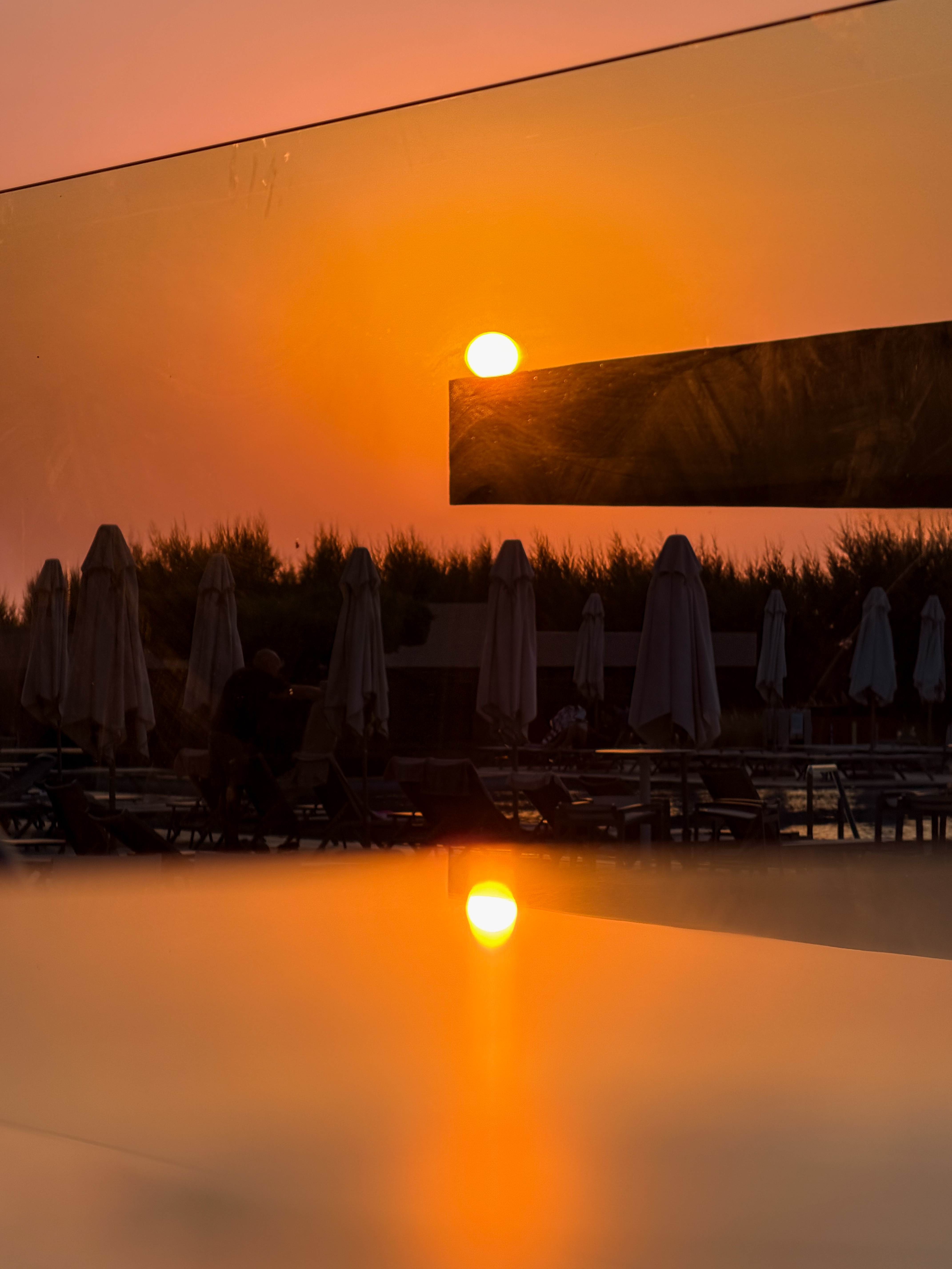 Bright orange sunrise over a pool with sun umbrellas, reflected in a smooth glass surface during early hours