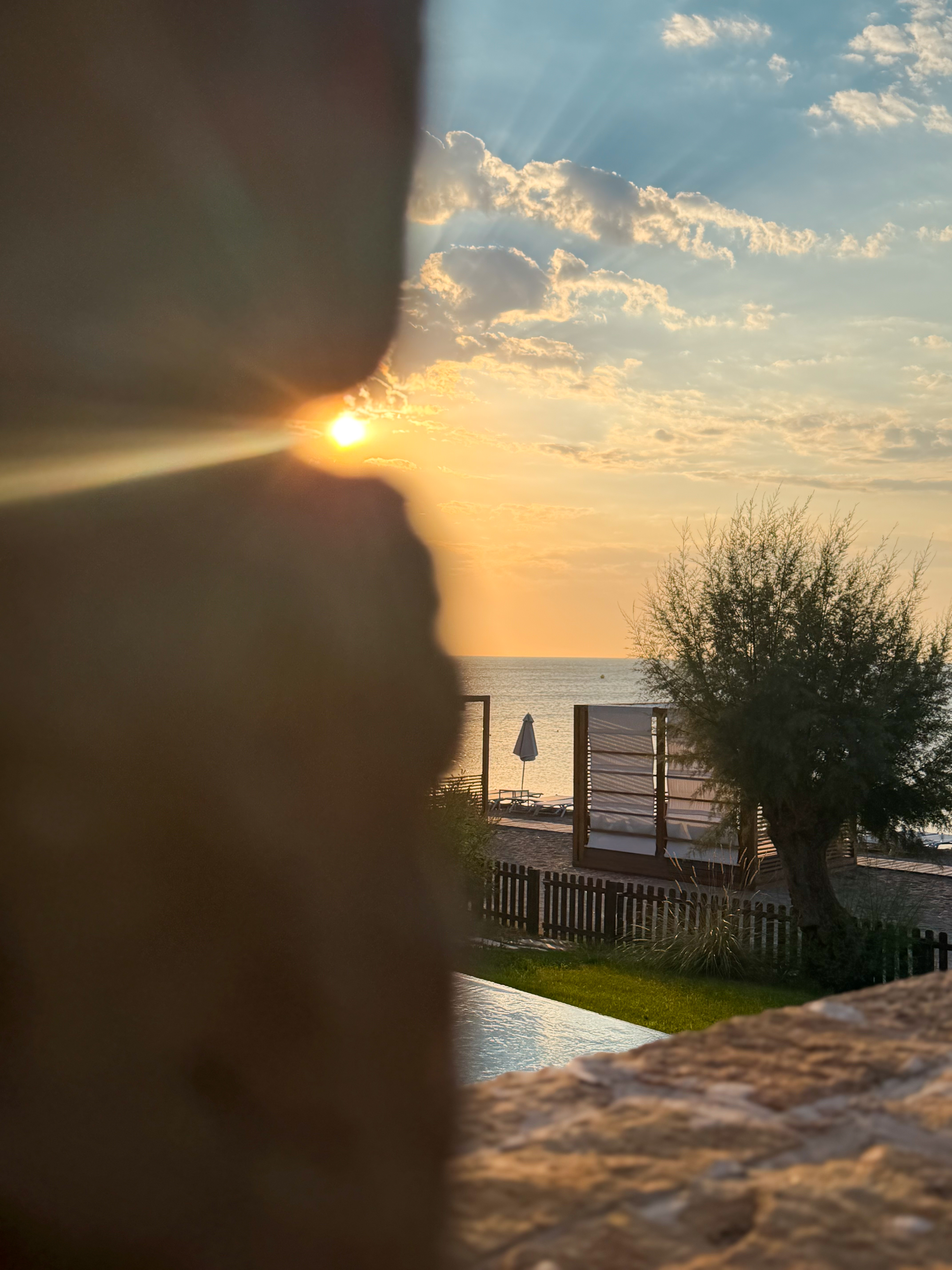 Golden sun rays peeking through a stone wall overlooking a quiet coastal garden and the sea at sunrise.