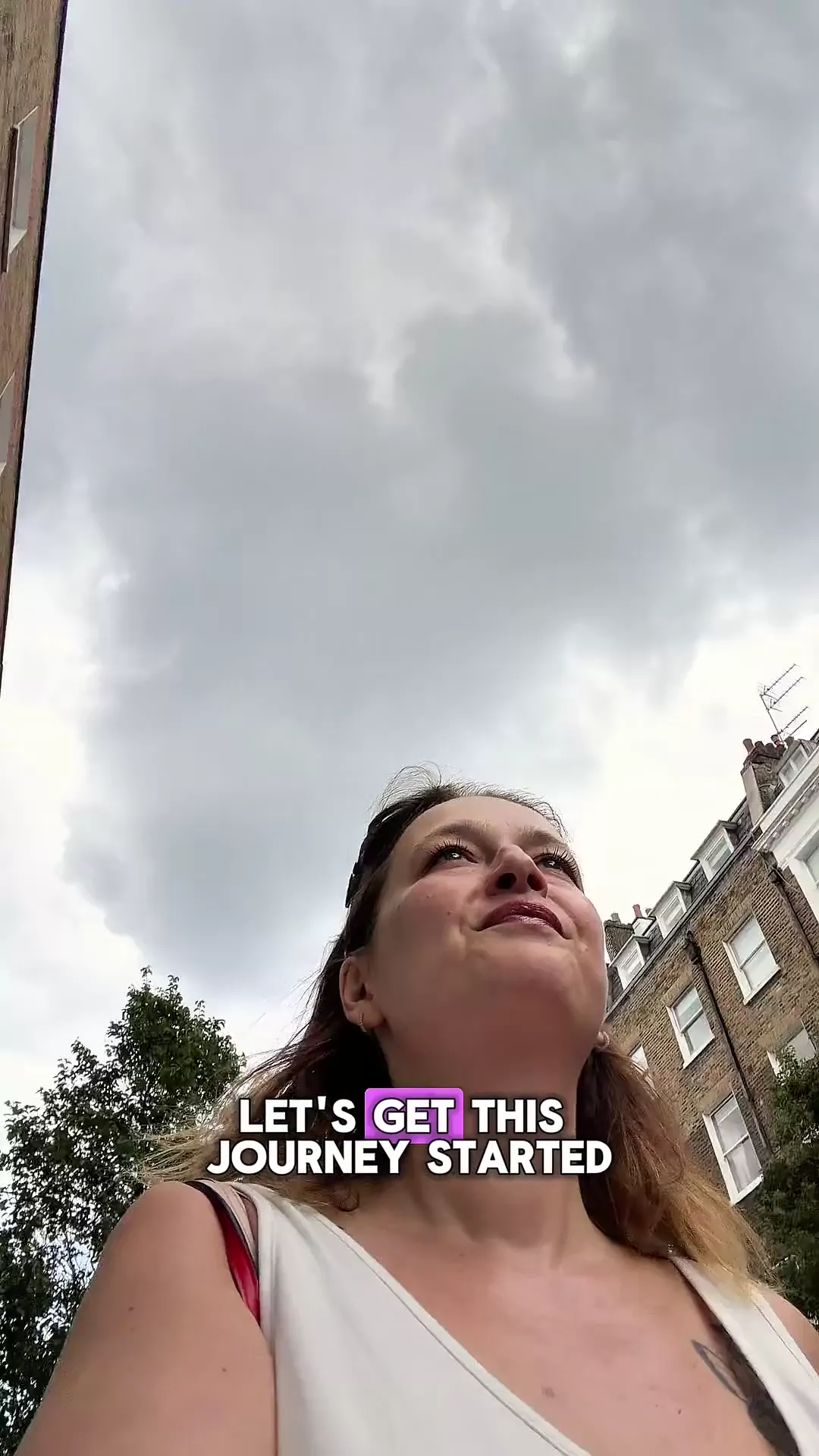 Low angle view of a woman looking up at a cloudy London sky, surrounded by brick buildings and greenery.
