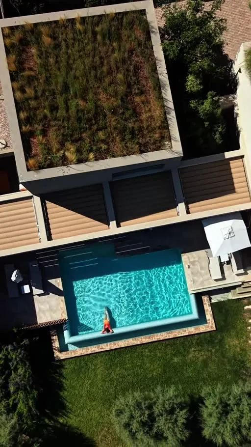Aerial view of a bright turquoise pool beside a grass-covered roof and geometric architecture in the midday sun.