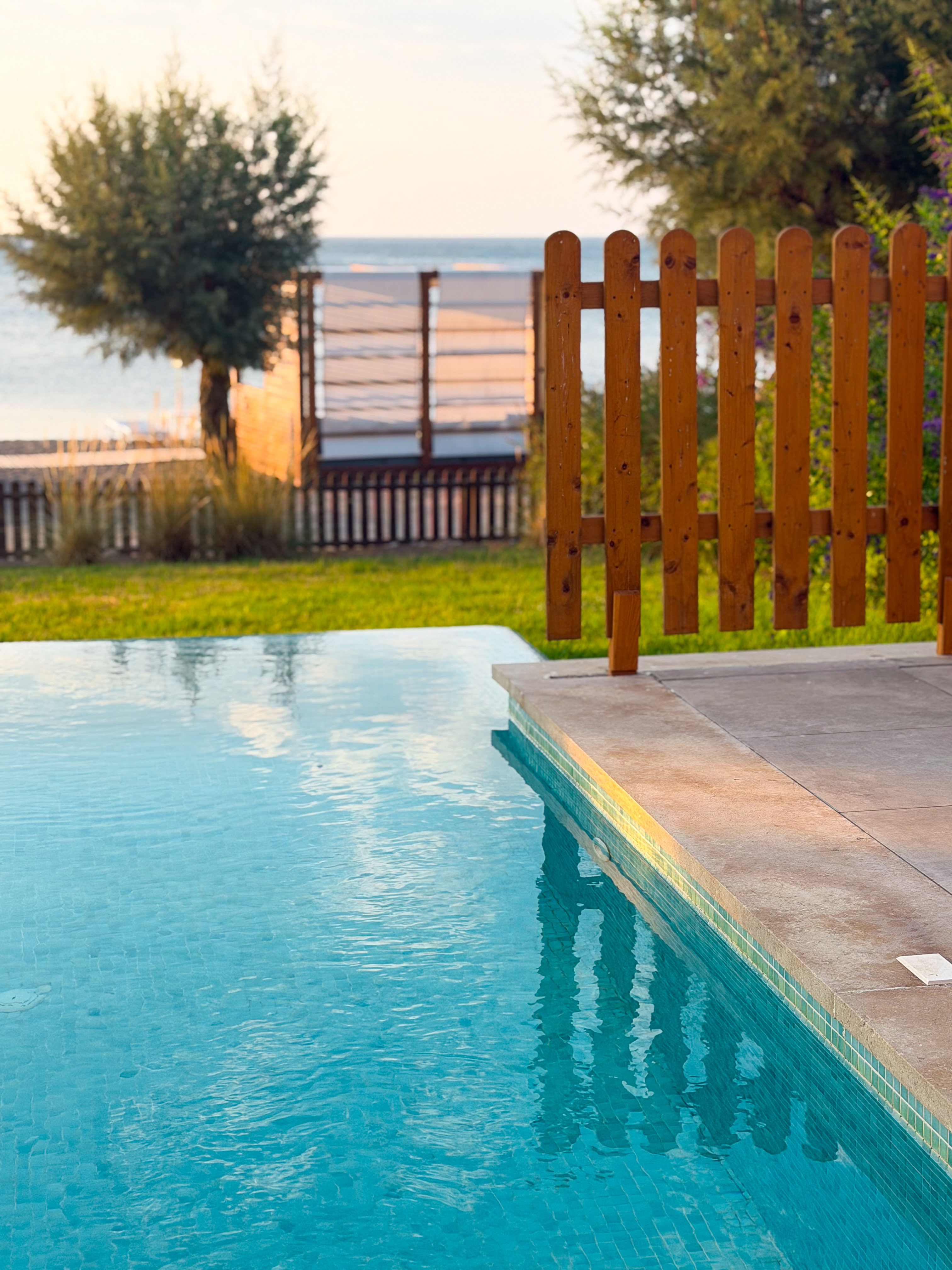 A blue tiled swimming pool corner next to a wooden fence with sea and trees in the soft evening light.