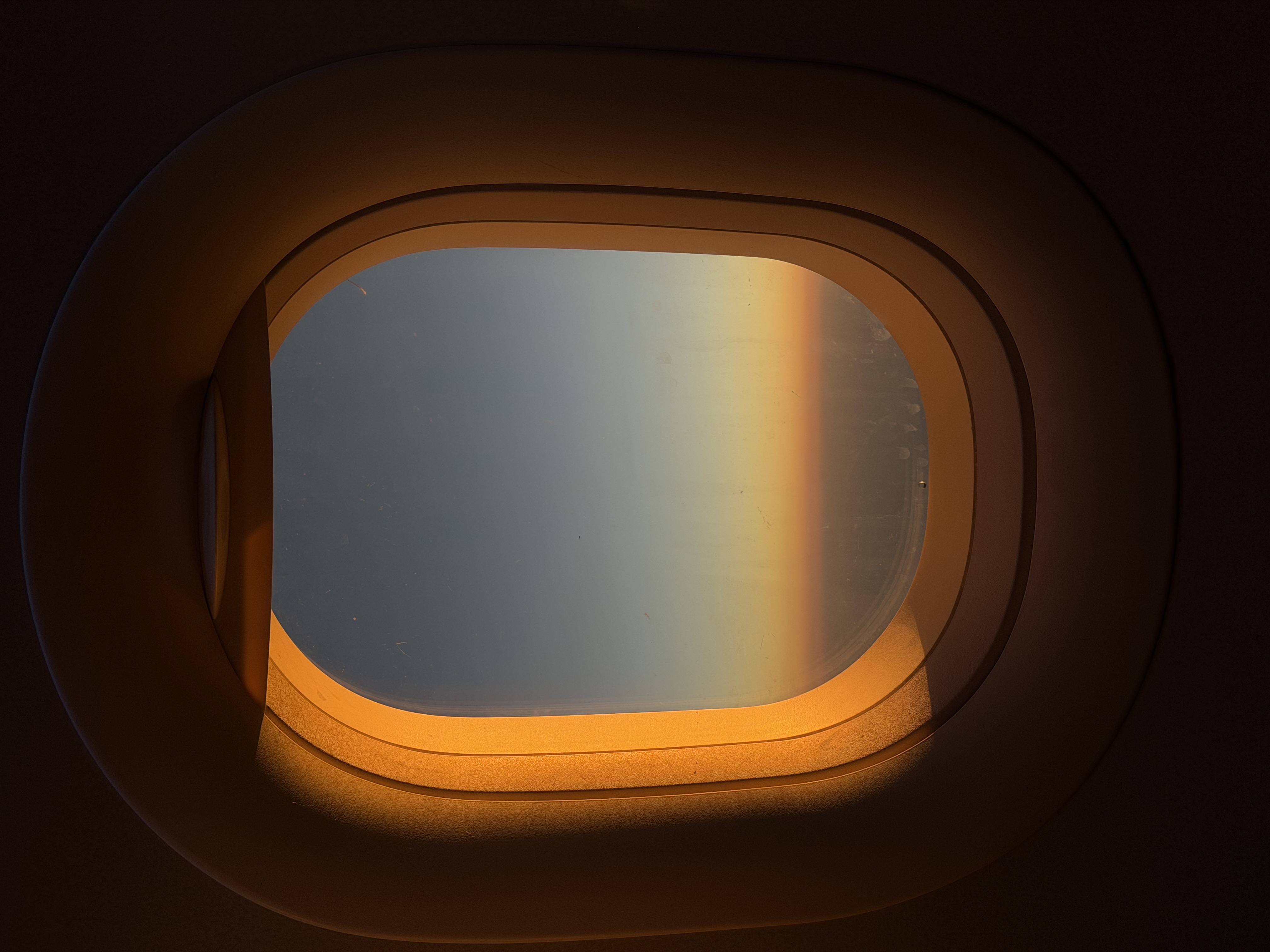 An airplane window frame captures a soft orange sunrise against a deep blue sky above the clouds. Warm, minimal tones.